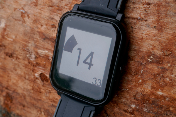 A smart watch displaying a speed gauge and large number "14" in a pretty font. It lies on a block of wood.